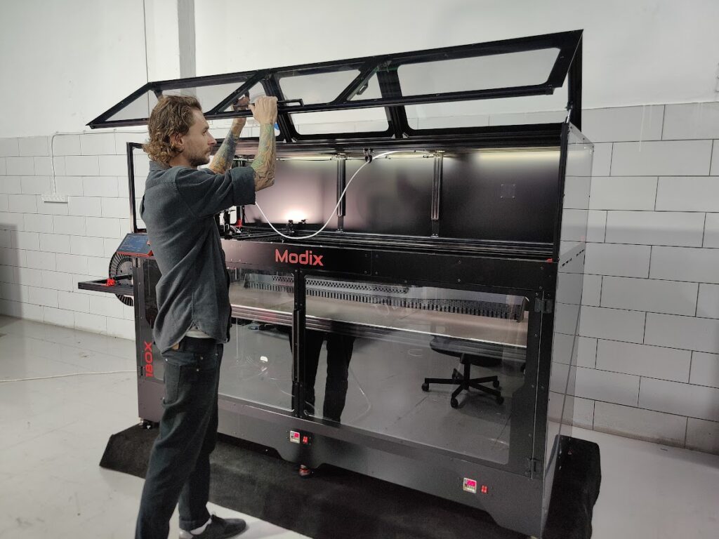 Imprimante 3D grand format Modix BIG-180X avec enceinte fermée, portes transparentes et capots supérieurs relevables pour faciliter l'accès au plateau d'impression.