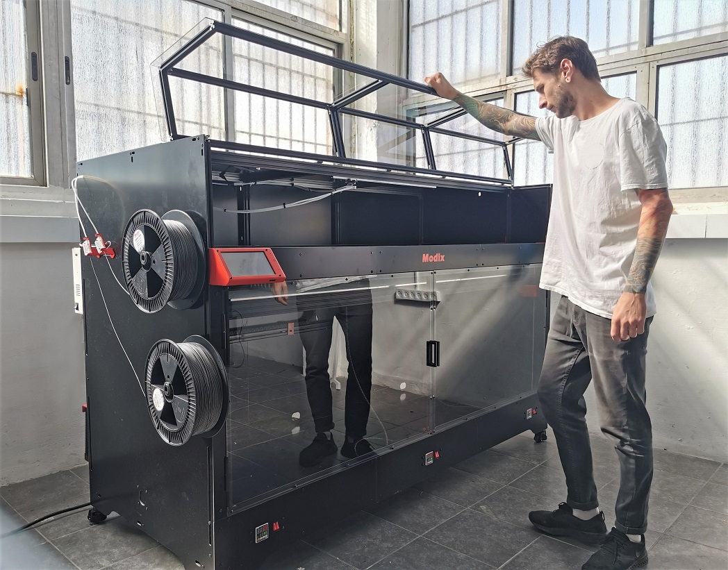 Imprimante 3D grand format Modix BIG-180X avec enceinte fermée et capot supérieur relevé pour faciliter l'accès au plateau d'impression.